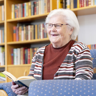 Eine lächelnde ältere Frau sitzt auf einem blauen Sessel und hält ein Buch in der Hand. Sie trägt eine gestreifte Strickjacke. Im Hintergrund sind Regale mit einer Vielzahl von Büchern zu sehen.