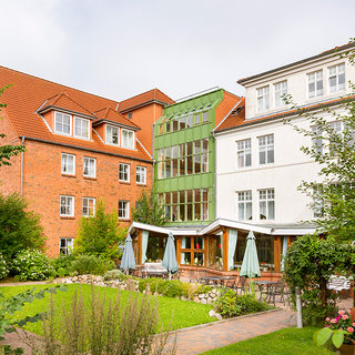 Ein Innenhof mit mehreren Gebäuden, darunter ein modernes, grünes Gebäude und traditionellere rote Ziegelbauten. Der Garten ist gepflegt, mit Bänken und Tischen, umgeben von Sträuchern und Bäumen. Schattenspendende Sonnenschirme sind aufgestellt, was eine einladende Atmosphäre schafft.