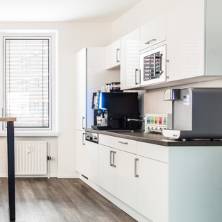Moderne Küche mit weißen Schränken und einem grauen Arbeitsbereich. An der Wand steht ein Kaffeeautomat, daneben verschiedene Kaffeekapseln. Ein Fenster mit Jalousien sorgt für Tageslicht. Holzboden und ein hoher Tisch runden das Bild ab.