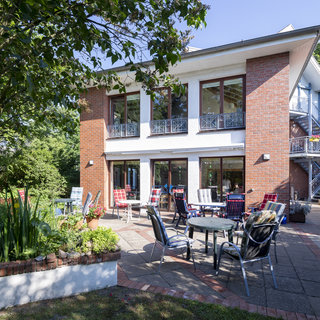 Ein zweigeschossiges, modern gestaltetes Haus mit roten Ziegeln. Vor dem Gebäude befindet sich ein gepflasterter Außenbereich mit Tischen und Stühlen. Umgeben von grünen Bäumen und Pflanzen, strahlt die Szenerie eine einladende und entspannte Atmosphäre aus.