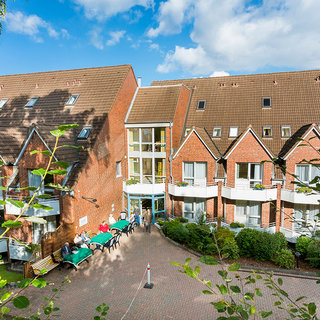 Eine gepflegte Unterkunft mit roten Ziegelbauten und einem einladenden Innenhof. Auf den Terrassen sitzen Gäste unter Sonnenschirmen. Die Umgebung ist grün mit Büschen und Bäumen. Der Himmel ist klar und blau, was eine freundliche Atmosphäre schafft.