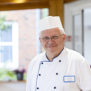 Ein lächelnder Koch trägt eine weiße Kochjacke und eine Kochmütze. Er steht in einer hellen Umgebung mit Pflanzen im Hintergrund. Der Koch vermittelt eine freundliche und einladende Atmosphäre.