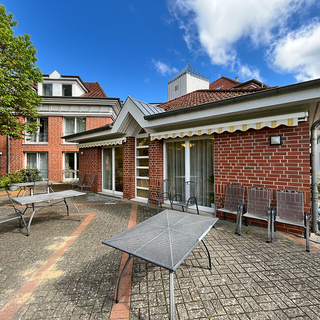 Eine Terrasse mit mehreren Tischen und Stühlen, umgeben von roten Backsteinwänden. Im Hintergrund sind Bäume und ein Gebäude zu sehen. Der Himmel ist blau mit einigen Wolken.