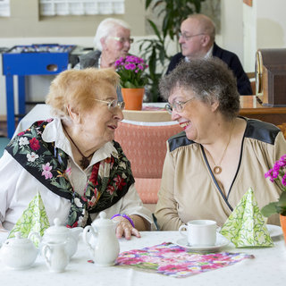 Zwei ältere Frauen sitzen an einem Tisch und lächeln einander an, während sie sich unterhalten. Auf dem Tisch stehen Teekannen und Blumen in Töpfen. Im Hintergrund sind weitere Personen zu sehen, die in einer Cafeteria verweilen.