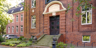 Die Ansicht zeigt den Eingang eines alten, roten Backsteingebäudes mit einer Treppe und einer markanten Eingangstür. Über der Tür befindet sich ein Schild mit der Aufschrift „Kaminhaus“. Die Umgebung ist von Bäumen und grünem Bewuchs geprägt.