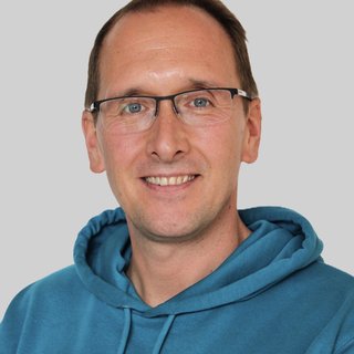 Eine lächelnde Person trägt einen blauen Kapuzenpullover mit dem Logo „CURA“. Die Person hat Brille und einen Namensschild, der auf den Namen „Daniel G. “ hinweist. Der Hintergrund ist grau, wodurch die Person klar hervorsticht.