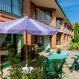 Im Bild ist eine sonnige Terrasse mit mehreren Gartenmöbeln zu sehen, darunter ein Tisch mit Stühlen und einem Sonnenschirm. Im Hintergrund befindet sich ein Gebäude mit Balkonen und Fenstern. Die Umgebung ist grün und einladend, geeignet für entspannte Stunden im Freien.