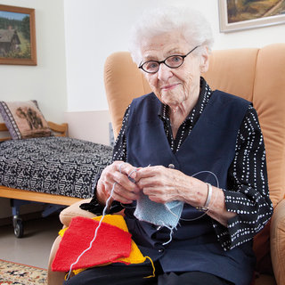Eine ältere Frau mit Brille sitzt in einem Sessel und strickt mit buntem Garn. Sie trägt eine schwarze Weste mit weißen Punkten und schaut freundlich. Im Hintergrund ist ein Bett und ein Bild an der Wand zu sehen.