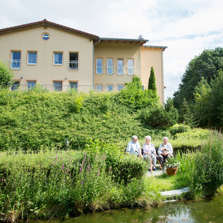 Drei ältere Menschen sitzen an einem kleinen Teich, umgeben von einer grünen Landschaft und blühenden Pflanzen. Im Hintergrund ist ein helles Gebäude sichtbar. Die Szene strahlt Ruhe und Gemeinschaft aus.