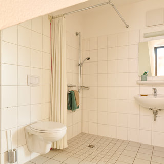 Das Bild zeigt ein helles Badezimmer mit weißen Fliesen. Links befindet sich eine Dusche mit einem Duschvorhang, rechts ein Waschbecken mit einem Spiegel darüber. Ein Toilettenbecken ist an der Wand befestigt, und Fensterlicht fällt in den Raum.