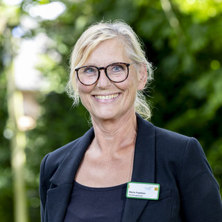 Eine lächelnde Frau mit blonden, lockigen Haaren trägt eine schwarze Blazerjacke und eine Brille. Sie steht vor einem verschwommenen, grünen Hintergrund und hat ein Namensschild befestigt, das ihren Namen zeigt. Die Atmosphäre ist freundlich und einladend.