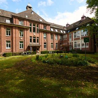 Ein historisches Gebäude mit rotem Backstein und großen Fenstern, umgeben von einem gepflegten Garten. Im Vordergrund sind Bäume und ein Sitzbereich mit Bänken zu sehen. Der Himmel ist teilweise bewölkt, und es herrscht eine ruhige, einladende Atmosphäre.