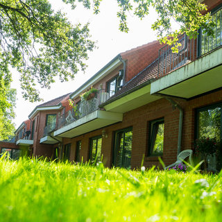 Das Bild zeigt eine Reihe von низких Gebäuden mit Balkonen, umgeben von grünem Gras und Bäumen. Es herrscht sonniges Wetter, was eine einladende und ruhige Atmosphäre vermittelt. Die Gebäude sind aus rotem Ziegelstein und verfügen über große Fenster.