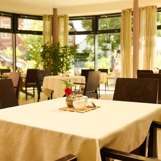 Ein helles Restaurant mit mehreren Tischen, die mit weißen Tischdecken bedeckt sind. In der Mitte eines Tisches steht eine kleine Vase mit einer roten Blume. Große Fenster lassen viel Tageslicht herein und bieten eine Sicht auf das Außenareal mit Bäumen und Menschen.