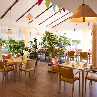 Ein helles, einladendes Restaurant mit mehreren Tischen und Stühlen, umgeben von Pflanzen. Die Wände sind in warmen Gelbtönen gehalten, und bunte Wimpel schmücken die Decke. Die große Fensterfront lässt Tageslicht herein und bietet einen Blick nach draußen.