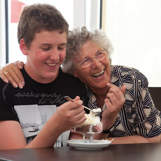 Ein lachender Jugendlicher und eine fröhliche ältere Frau sitzen an einem Tisch. Sie genießen Eiscreme und teilen einen glücklichen Moment miteinander. Die Frau legt ihre Hand nachdenklich auf die Schulter des Jungen, was eine enge Beziehung zwischen ihnen zeigt.