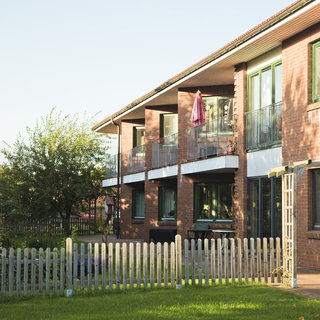 Ein zweigeschossiges Backsteingebäude mit Balkonen, auf denen eine lila Markise hängt. Vor dem Gebäude befindet sich ein Zaun aus Holzlatten, und im Hintergrund sind einige Bäume zu sehen. Der Rasen ist grün und gepflegt.