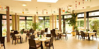 Ein heller Restaurantbereich mit Tischdecken und Stühlen. Einige Tische sind für Gäste gedeckt, mit Wasserflaschen und Tellern. Große Fenster lassen Tageslicht herein und bieten Ausblick auf einen Garten. An der Decke hängen dekorative Elemente aus Papier.