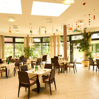 Ein heller Restaurantbereich mit Tischdecken und Stühlen. Einige Tische sind für Gäste gedeckt, mit Wasserflaschen und Tellern. Große Fenster lassen Tageslicht herein und bieten Ausblick auf einen Garten. An der Decke hängen dekorative Elemente aus Papier.