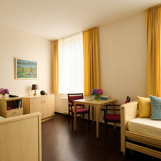 Der Raum zeigt eine helle, freundliche Einrichtung mit zwei Betten, einem Tisch und Stühlen. Große Fenster mit gelben Vorhängen ermöglichen viel Tageslicht. An der Wand hängt ein Gemälde, und auf dem Tisch stehen Blumen in einer Vase.