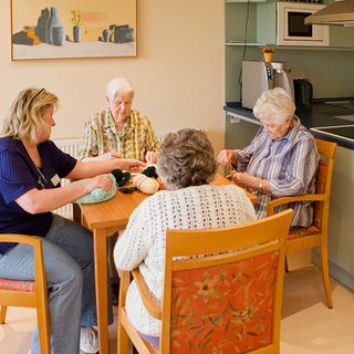 Vier Frauen sitzen um einen Tisch in einer hellen Innenräumlichkeit. Eine ältere Frau strickt, während die anderen beobachten und sich unterhalten. Im Hintergrund befindet sich eine Küche mit modernen Geräten. Die Atmosphäre ist freundlich und gemeinschaftlich.