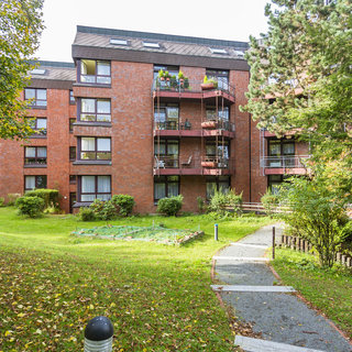Ein mehrstöckiges, rotes Mauerwerk-Gebäude mit Balkonen ist von grünen Wiesen und Bäumen umgeben. Ein gepflasterter Weg führt durch die Landschaft. Die Fenster sind hell und der Zugang ist übersichtlich gestaltet. Es herrscht eine ruhige, einladende Atmosphäre.