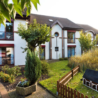 Ein zweigeschossiges, helles Wohnhaus mit mehreren Fenstern und rotem Rolladen. Vor dem Haus befindet sich ein kleiner Garten mit Bäumen, Büschen und einem eingezäunten Bereich, der ein kleines Spielhaus enthält. Die Atmosphäre ist freundlich und einladend.