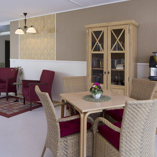 Ein gemütlicher, heller Raum mit einem Tisch und vier Stühlen, umgeben von einem Bücherregal und einer Ecke mit zwei roten Sesseln. Ein dekorativer Teppich verleiht dem Raum eine einladende Atmosphäre. Ein Getränkeautomat steht in der Nähe.