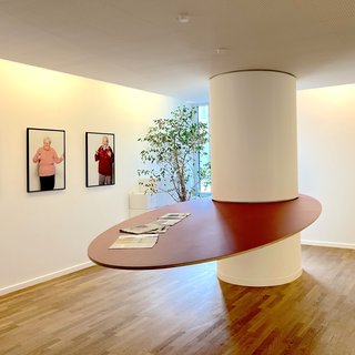 Der Raum zeigt eine moderne Innenarchitektur mit einem runden Tisch und mehreren gerahmten Porträtfotos älterer Menschen an der Wand. Ein grünes Pflanzelement ergänzt die freundliche Atmosphäre. Der Boden ist aus Holz und verleiht dem Raum Wärme.