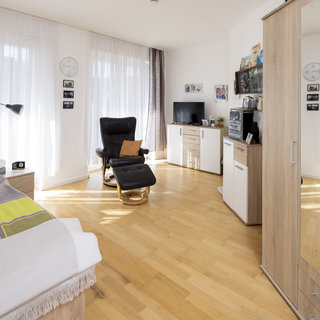 Ein helles Schlafzimmer mit Holzboden, ausgestattet mit einem Bett, einem schwarzen Relaxstuhl und einem TV-Schrank. Große Fenster mit weißen Vorhängen lassen viel Licht herein. An der Wand hängen Fotos, und eine Uhr zeigt die Zeit an. Die Einrichtung ist modern und gemütlich.