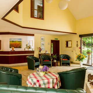 Ein Empfangsbereich mit einer hellen, freundlichen Atmosphäre. Im Vordergrund stehen grüne Sessel um einen Tisch mit einem karierten Tischtuch. Im Hintergrund ist ein Empfangstresen zu sehen, sowie ein großer Fensterbereich mit Blick auf Pflanzen und Fahrräder im Freien.