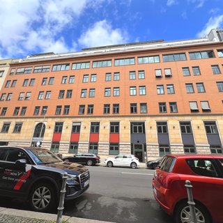 Eine moderne, fünfstöckige Backsteingebäude-Fassade mit großen Fenstern. Auf der Straße parken mehrere Autos, darunter ein schwarzer und ein roter Wagen. Über dem Gebäude ist ein blauer Himmel mit vereinzelten Wolken zu sehen.