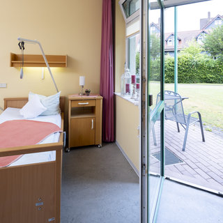 Ein heller, freundlicher Raum mit einem Krankenhausbett, einem Nachttisch und einem Fenster, das auf einen Garten blickt. Die Tür ist offen und führt auf einen gepflasterten Außenbereich mit einem Stuhl. Der Raum ist mit warmen Farben und einer weichen Beleuchtung gestaltet.