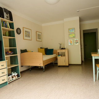 Das Bild zeigt einen geräumigen Raum mit einem Bett, einem Tisch und Stühlen. An den Wänden hängen Bilder und es gibt eine TV-Einheit. Im Hintergrund ist eine Tür sichtbar, die in einen weiteren Bereich führt. Der Raum wirkt freundlich und hell.