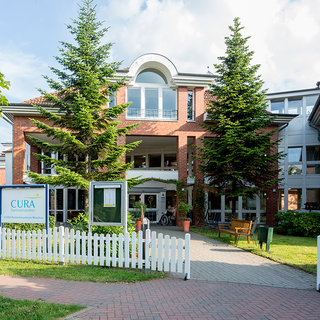 Ein rotes Backsteingebäude mit großen Fenstern und einem Eingangsbereich ist von grünen Bäumen umgeben. Vor dem Gebäude steht ein Schild mit der Aufschrift „CURA“. Im Vordergrund gibt es einen gepflasterten Weg und eine weiße Zaunumrandung.