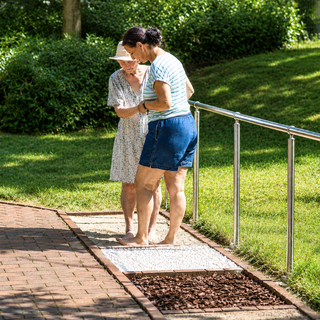 Zwei Frauen stehen barfuß auf einem speziellen Gehweg im Freien, umgeben von Rasen und Bäumen. Eine Frau in einem blauen Oberteil und Shorts erklärt etwas, während die andere Frau in einem blumigen Kleid interessiert zuhört.