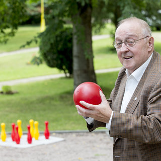 Ein älterer Mann mit Brille hält eine rote Kugel in seiner Hand und lächelt. Im Hintergrund stehen bunte Kegel (gelb und rot) auf einer Wiese. Es scheint, als würde er sich darauf vorbereiten, die Kugel zu werfen.