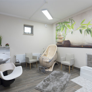 Ein heller, moderner Raum mit einem Waschbecken für Haarschnitte und einem bequemen Friseurstuhl. An der Wand ist ein Bild mit Pflanzen und Steinen. Es gibt mehrere Stühle und einen Teppich aus Grau, der dem Raum eine einladende Atmosphäre verleiht.