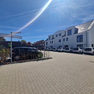 Eine Straße mit modernen, weißen Wohngebäuden auf der rechten Seite. In der Mitte steht ein Wegweiser mit den Beschriftungen „WohnGemeinschaft“, „PflegeZuG“ und „ServiceWohnungen“. Links ist ein Parkplatz mit mehreren Autos, die geparkt sind. Der Himmel ist klar und sonnig.