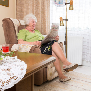 Eine ältere Dame sitzt bequem in einem Sessel und liest eine Zeitung. Sie trägt ein grünes Oberteil und einen Blumenrock. Vor ihr steht ein Tisch mit einem Glas Getränk. Die Szene strahlt Gemütlichkeit und Entspannung aus.