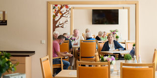 Eine Gruppe älterer Menschen sitzt in einem hellen Raum mit Tischen und Stühlen, umgeben von Pflanzen. Einige Personen unterhalten sich, während andere an ihrem Essen beschäftigt sind. An der Wand hängt ein Fernseher. Der Raum strahlt eine freundliche Atmosphäre aus.