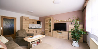 Ein helles Wohnzimmer mit einem großen, bequemen Sofa, einem Couchtisch und einem modernen Fernseher. An der Wand befinden sich eine Holzkommode und Regale mit dekorativen Objekten. Eine Pflanze steht in der Ecke, und Fenster mit Vorhängen lassen viel Licht herein.