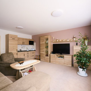 Ein helles Wohnzimmer mit einem großen, bequemen Sofa, einem Couchtisch und einem modernen Fernseher. An der Wand befinden sich eine Holzkommode und Regale mit dekorativen Objekten. Eine Pflanze steht in der Ecke, und Fenster mit Vorhängen lassen viel Licht herein.