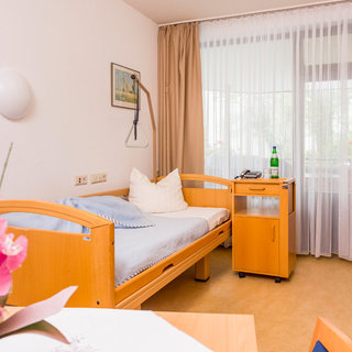 Ein helles Zimmer mit einem Pflegebett aus Holz, kissenverziert und einem kleinen Tisch daneben. Eine Flasche Wasser steht auf einem Rollwagen, und große Fenster mit Vorhängen lassen viel Tageslicht herein. Eine Blume ist im Vordergrund sichtbar.