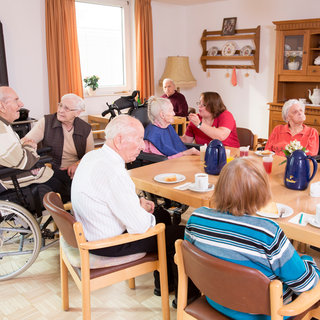 Eine Gruppe älterer Menschen sitzt in einem hellen Raum zusammen. Einige unterhalten sich, während andere beschäftigt sind, Getränke oder Snacks zu genießen. Es sind verschiedene Stühle und Tische vorhanden, und insgesamt herrscht eine gesellige Atmosphäre.