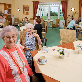 Im Bild ist ein hell erleuchteter Raum eines Gemeinschaftsbereichs zu sehen. Mehrere ältere Menschen sitzen an Tischen, trinken Kaffee und unterhalten sich. Einige sind in freundlicher Kleidung, und die Atmosphäre wirkt einladend und gesellig. Blühende Pflanzen sind auf den Tischen dekoriert.