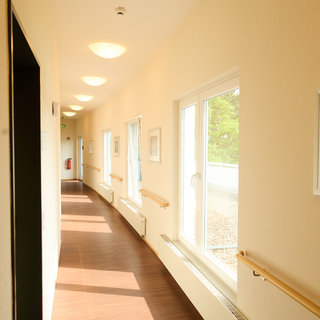 Eine helle Fluransicht mit durchgehenden Fenstern, die für Tageslicht sorgen. An den Wänden hängen Bilder, und der Boden ist aus Holz. Handläufe sind installiert, was auf eine barrierefreie Gestaltung hinweist.