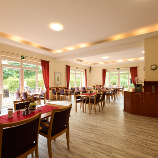 Ein helles Restaurant mit Tischen und Stühlen, gedeckt mit roten Tischläufern. Große Fenster lassen natürliches Licht herein und bieten Blick auf einen Garten. Der Raum ist stilvoll eingerichtet, mit rot Vorhängen und einer Servicetheke im Hintergrund.