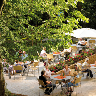 Eine Gruppe von Menschen sitzt an Tischen im Freien, umgeben von Bäumen und Blumen. Einige Personen nutzen Rollstühle. Es herrscht eine entspannte Atmosphäre, in der die Anwesenden Getränke genießen und miteinander sprechen. Ein Sonnenschirm bietet Schatten.