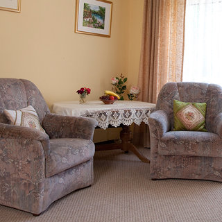 Zwei bequeme Sessel stehen in einem hellen Raum gegenüber. Zwischen ihnen befindet sich ein Tisch mit einer Spitze. Auf dem Tisch stehen Blumen und eine Schale mit Früchten. Die Wände sind in warmen Farben gehalten und es gibt Vorhänge, die Licht hereinlassen.